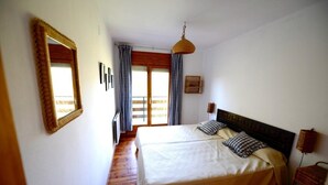 A bedroom with a large bed, a wooden floor, a hanging light fixture, and a window with curtains.