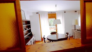 A room with a wooden floor, a round table, a chair, a lamp, and a shelf with dishes.