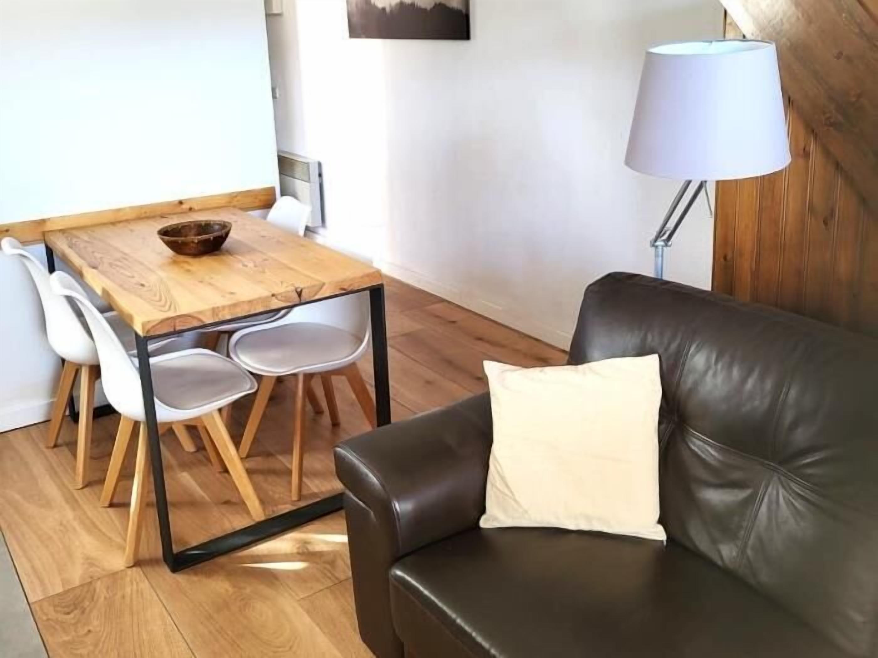 Ein gemütlicher Wohnbereich mit einem Holzesstisch, weißen Stühlen, einem schwarzen Ledersofa mit Kissen und einer Stehlampe.
