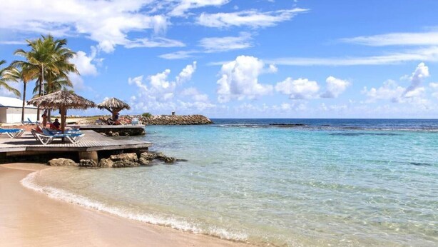 Una playa con agua turquesa cristalina, un muelle de madera con sillones de descanso y palmeras.