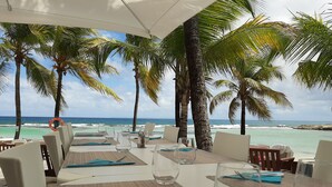 Un espacio para comer al aire libre con mesas dispuestas para dos, con vistas a una playa con palmeras y el océano.