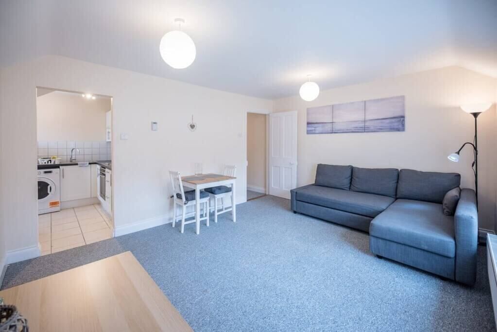 A modern living room with a grey sofa, a dining table with chairs, and a kitchen area visible through an open doorway.