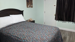 A bed with patterned bedding, a nightstand with a telephone, a door with a curtain, and a wall with a painting. - Laurel Notch Motel (Bordentown)