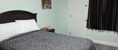 Una cama con ropa de cama estampada, una mesita de noche con teléfono, una puerta con cortina y una pared con un cuadro.