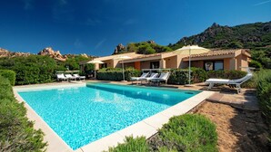 Une piscine entourée de chaises longues et de parasols, bordée de verdure et avec une chaîne de montagnes en toile de fond.