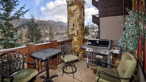 A balcony with a stone pillar, a grill, two chairs, and a table. - Beaver Creek Landing Condo 202 (Avon)