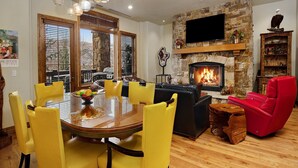 A living room with a stone fireplace, a round dining table with yellow chairs, a red armchair, and a television mounted on the wall. - Beaver Creek Landing Condo 202 (Avon)