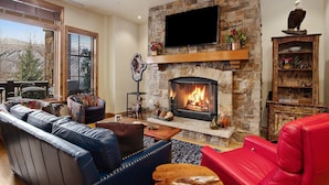 A cozy living room with a stone fireplace, leather sofas, a red chair, and a television mounted above the fireplace. - Beaver Creek Landing Condo 202 (Avon)