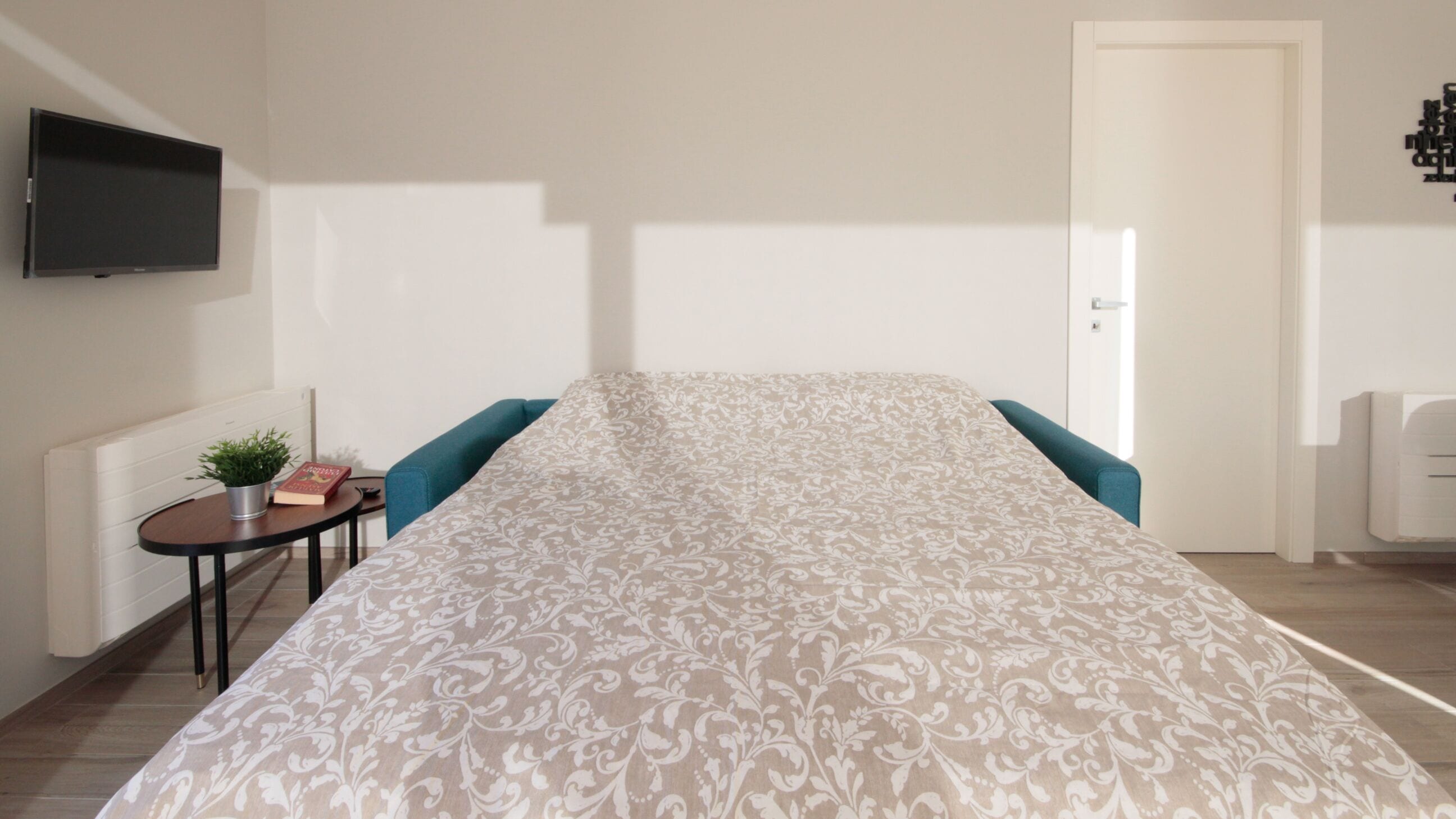 Une chambre moderne avec un grand lit, un téléviseur à écran plat fixé au mur et une petite table sur laquelle sont posées une plante et des livres.
