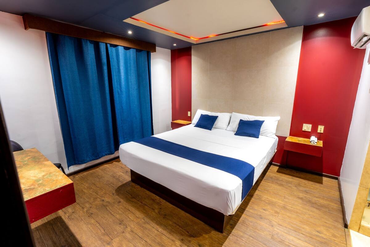 A modern hotel room with a large bed, wooden flooring, a red accent wall, and a blue curtain.