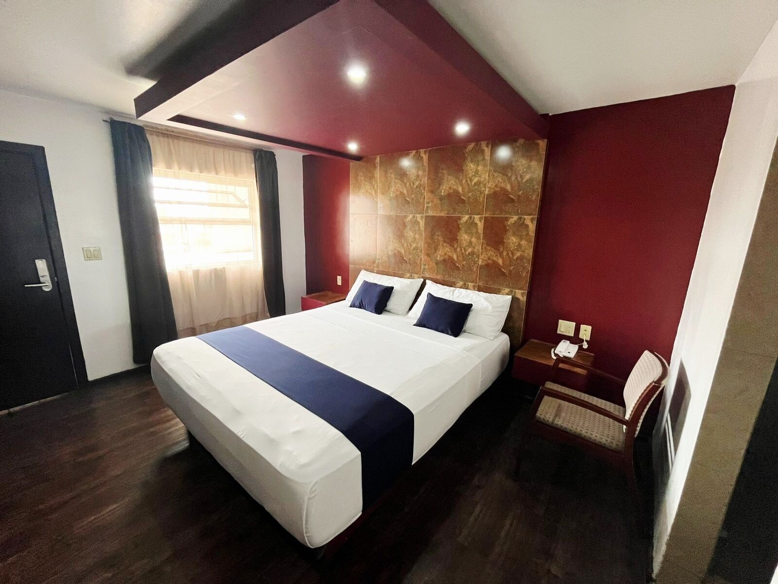 A hotel room with a large bed, a wooden headboard, a bedside table with a lamp, and a window with curtains.