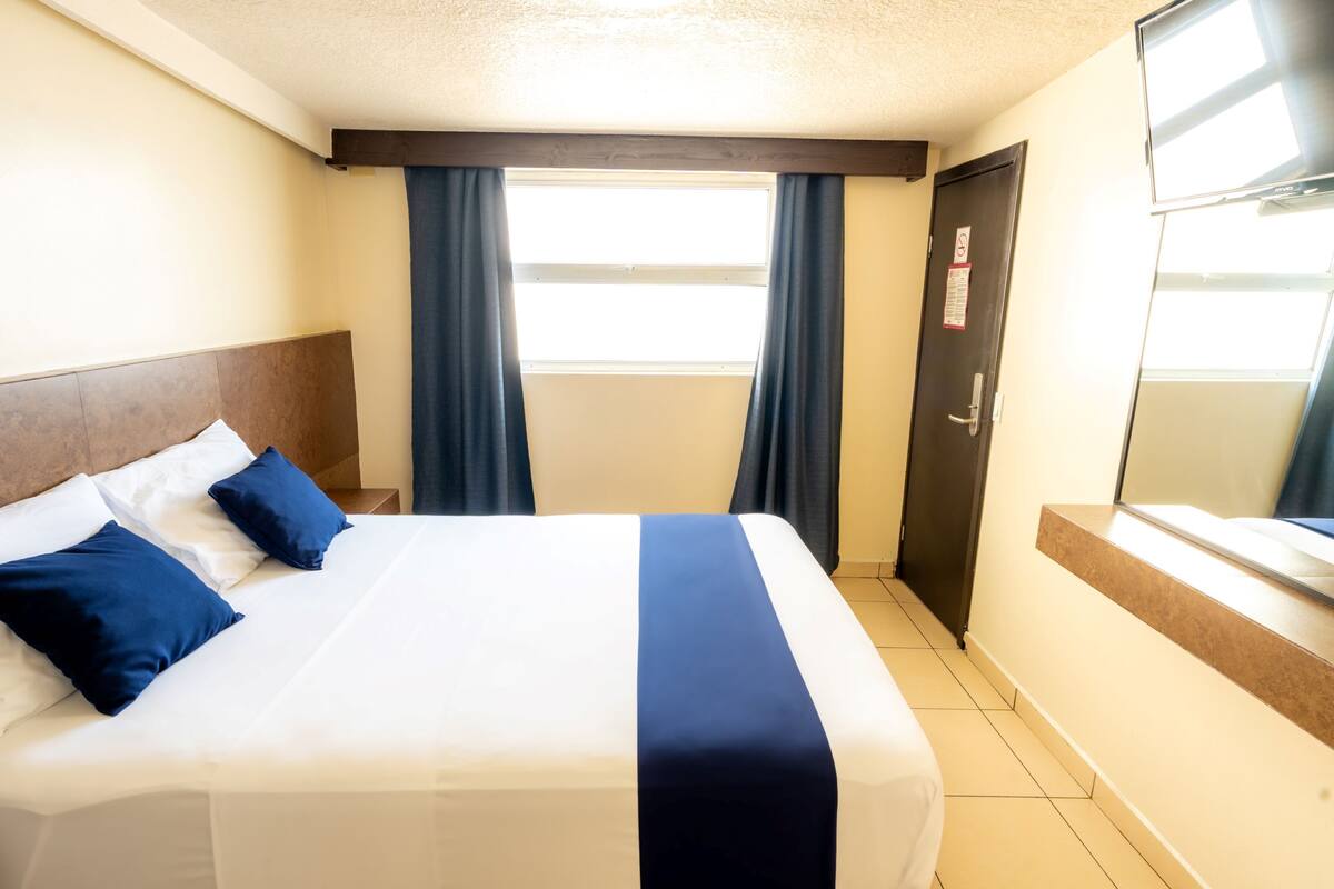A hotel room with a large bed, dark curtains, a door with a sign, and a window with blinds.