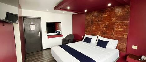 A hotel room with a bed, a desk, a chair, a TV, and a red accent wall.