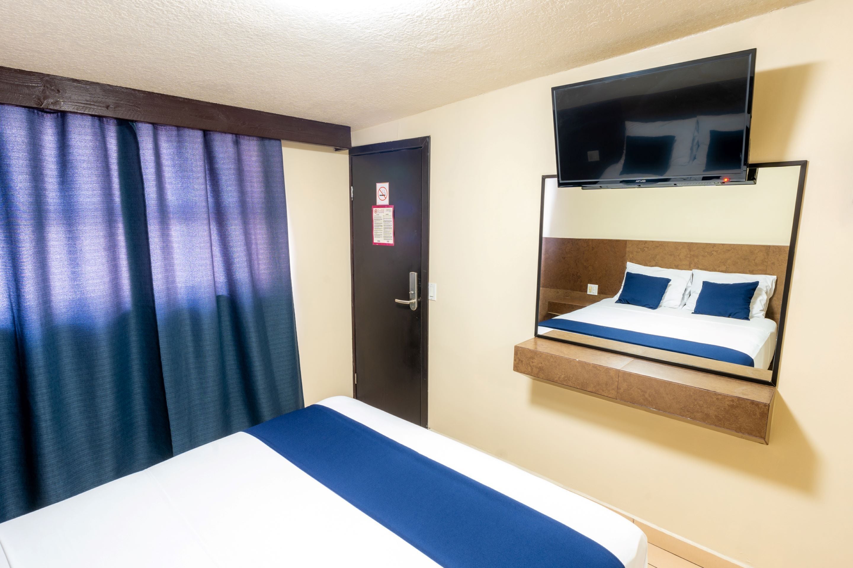 A hotel room with a bed, a mounted TV, and a door with a no-smoking sign.