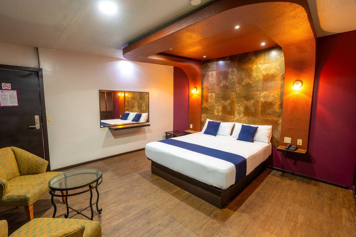 A hotel room with a large bed, a glass table, a chair, a desk, and a mirror.