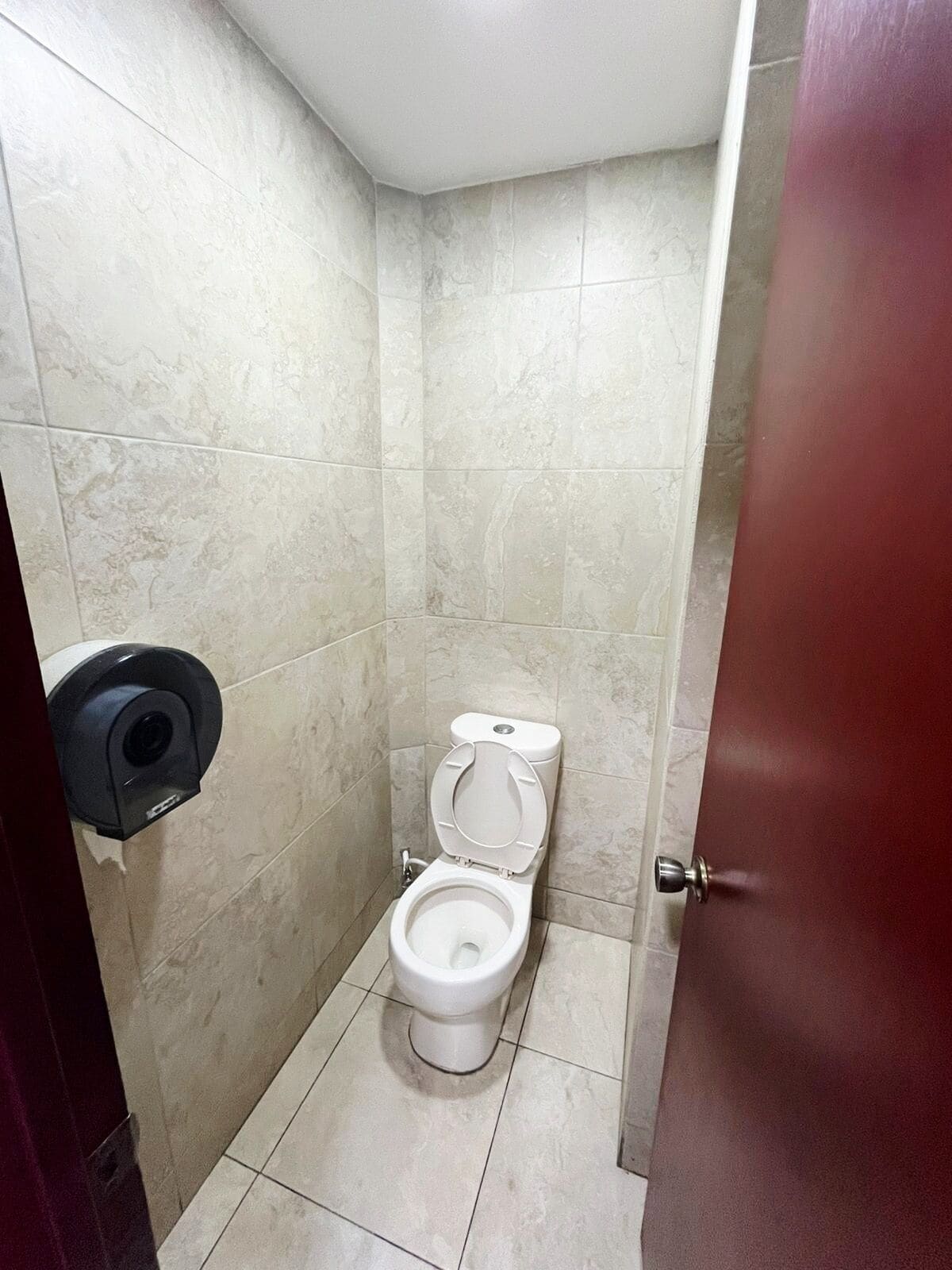 A small, tiled bathroom with a toilet, a paper towel dispenser, and a door.