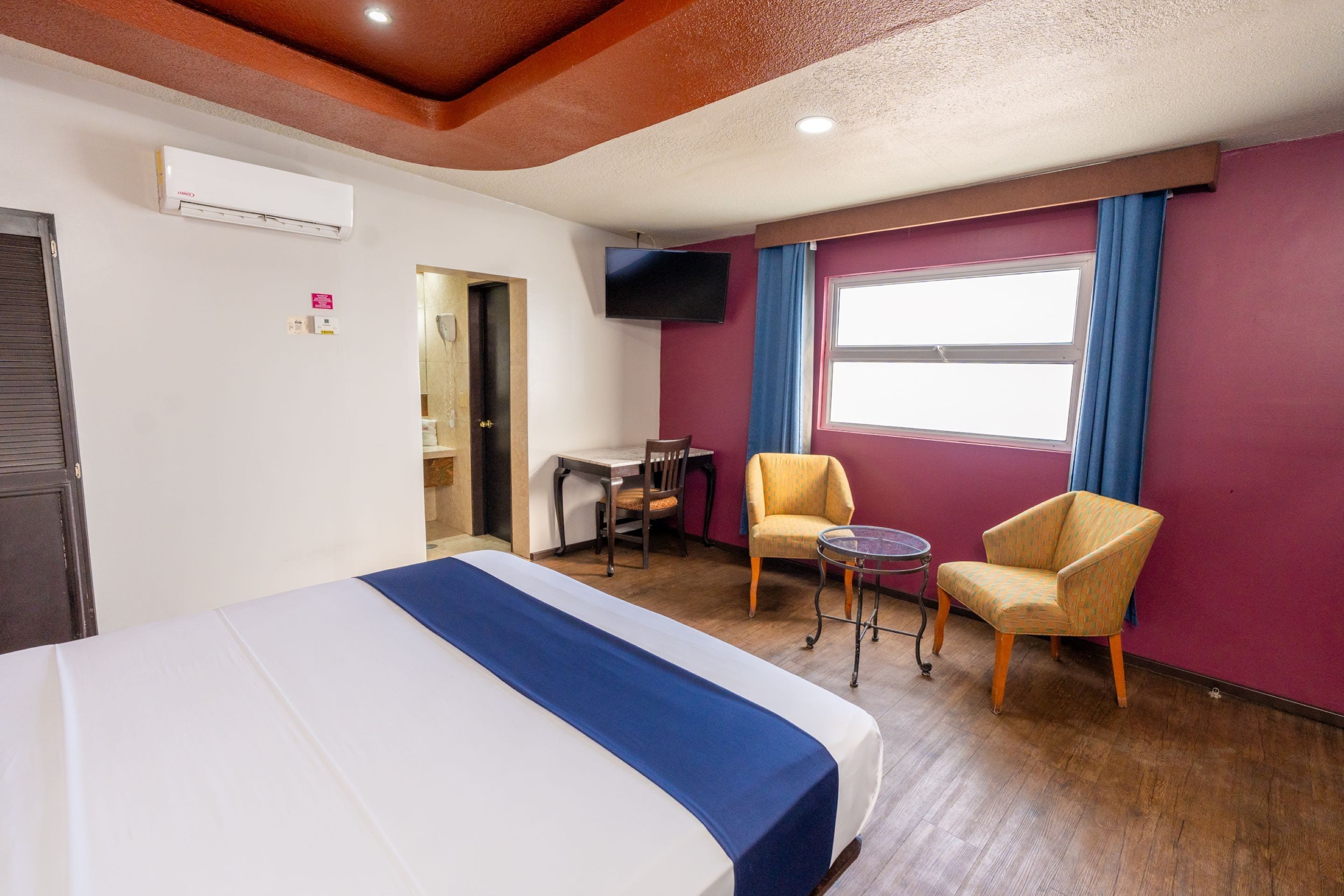 A hotel room with a bed, two armchairs, a small table, a TV, and a window with curtains.