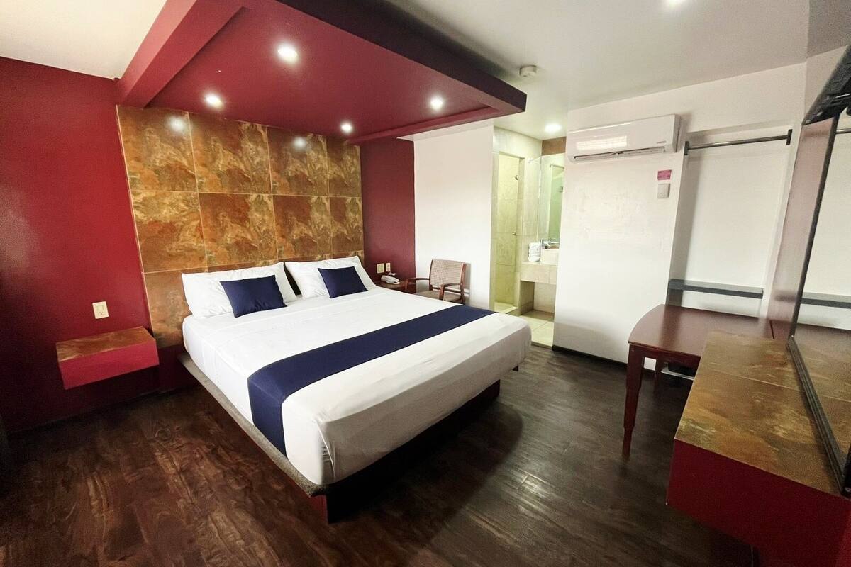 A hotel room with a large bed, a wooden floor, a red accent wall, and a bathroom with a shower and toilet.