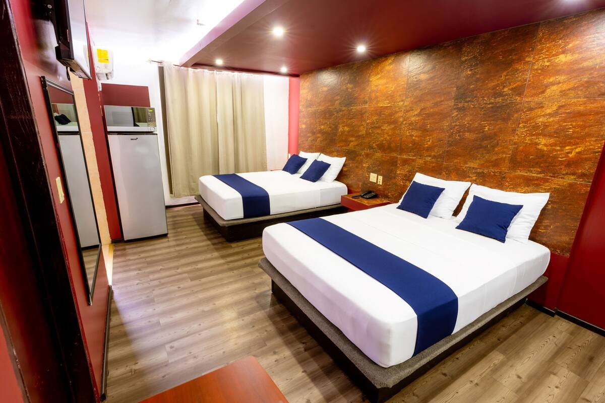 A hotel room with two beds, a wooden floor, a red accent wall, and a kitchenette.