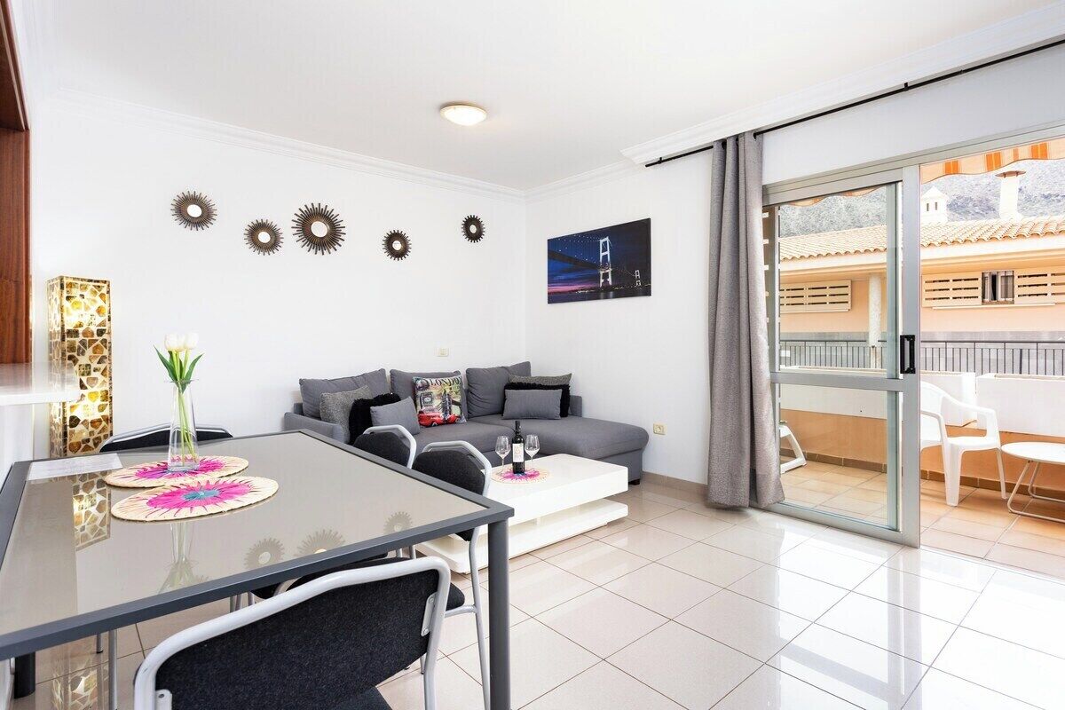 Un salon moderne avec une table à manger en verre, un canapé gris et un balcon agrémenté de chaises blanches.