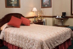 A neatly made bed with a patterned bedspread and two pillows, a bedside table with a lamp and a small plant, and framed pictures on the wall. - Britannia (San José)