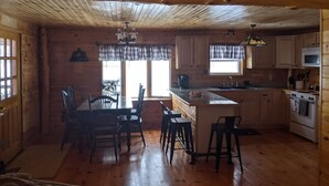 A wooden cabin interior with a dining area, kitchen, and large windows. - Knotty Pine Home- 2 Min Walk to Lake and Dock (Rangeley)