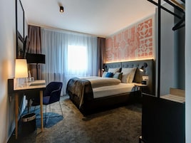 Una habitación de hotel con cama, escritorio, silla y un ventanal con cortinas.