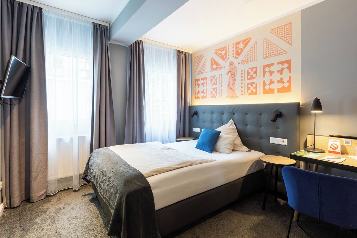 Hotellihuone, jossa on sänky, työpöytä, tuoli, televisio ja seinä, jossa on geometrisia kuvioita.