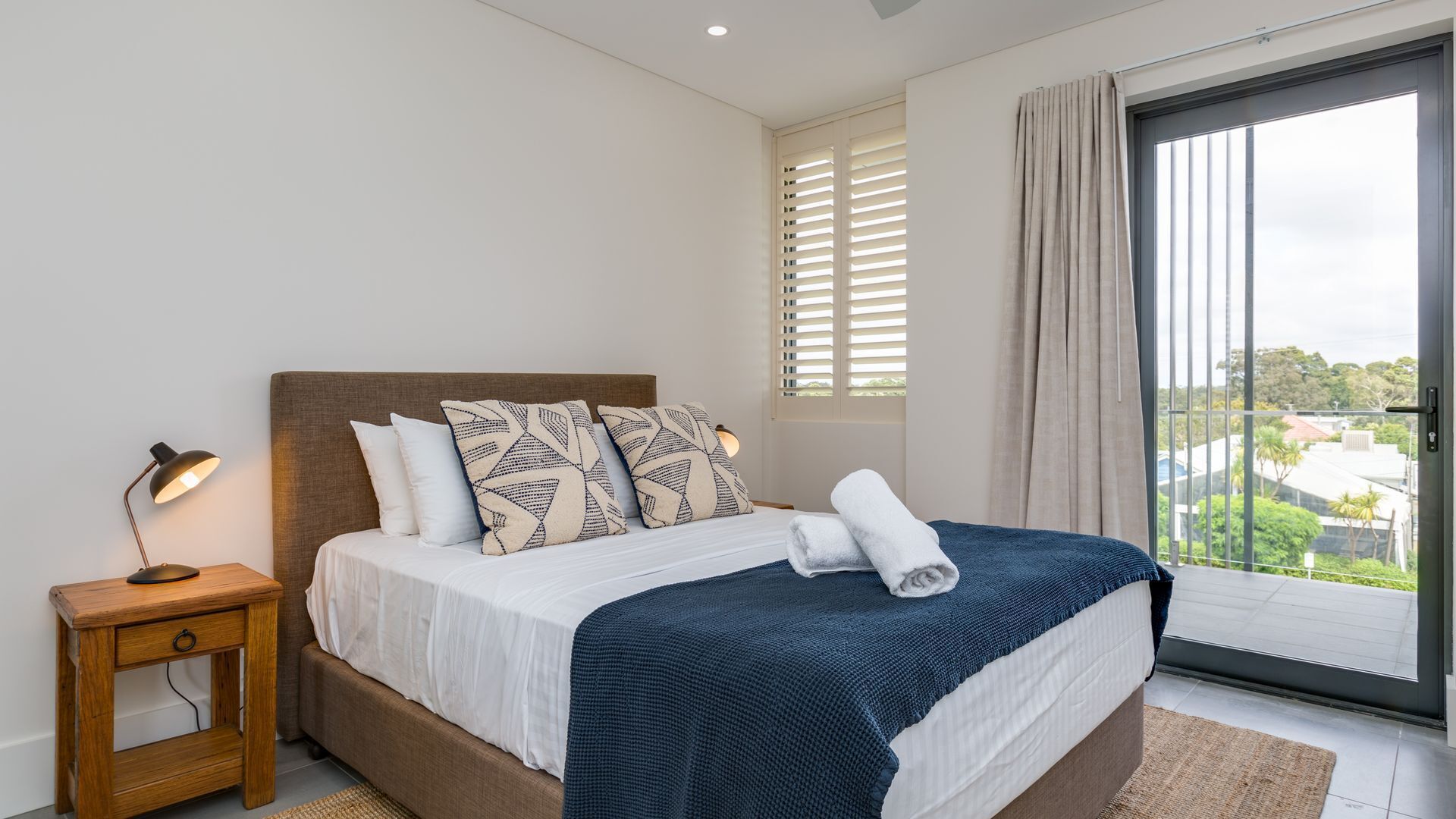 A bedroom with a bed, a nightstand, a lamp, a window with blinds, and a view of the outside.