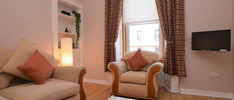Una sala de estar con un sofá color beige, un sillón a juego, una mesa de centro de vidrio con un control remoto, y un televisor en la pared.
