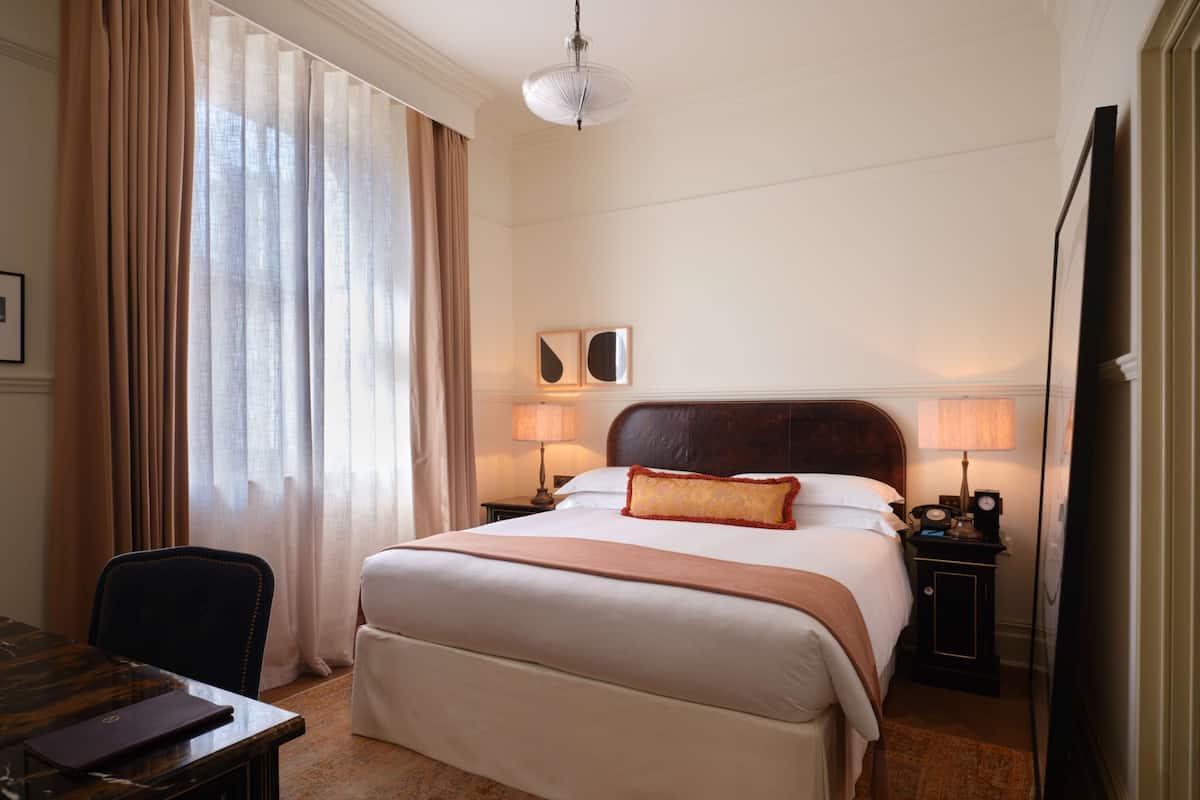 Quarto de hotel com uma cama grande, duas luminárias de cabeceira, uma escrivaninha com um laptop e uma cadeira.