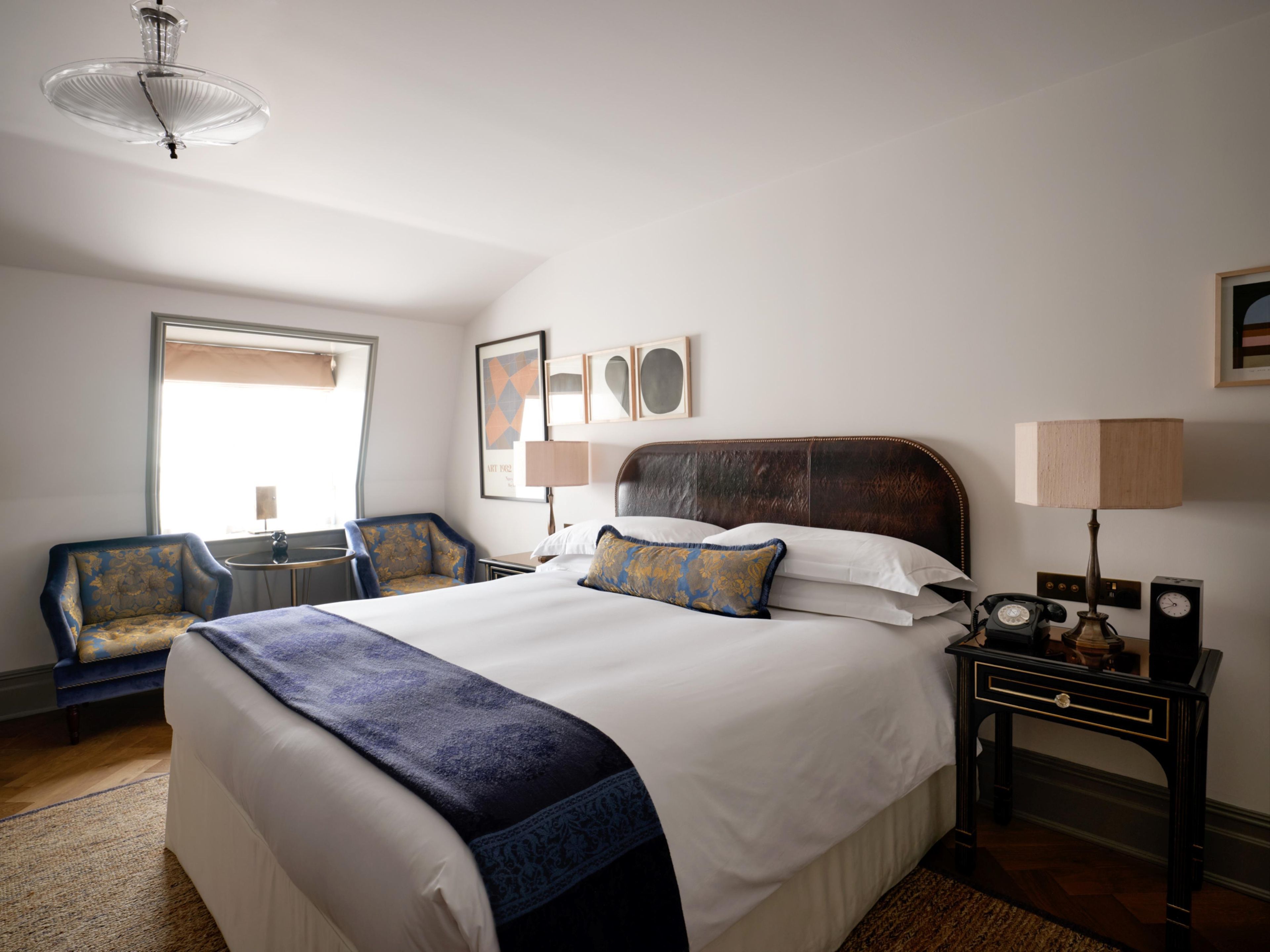 A hotel room with a large bed, a sitting area with a chair, a desk with a lamp, and a wall-mounted telephone.