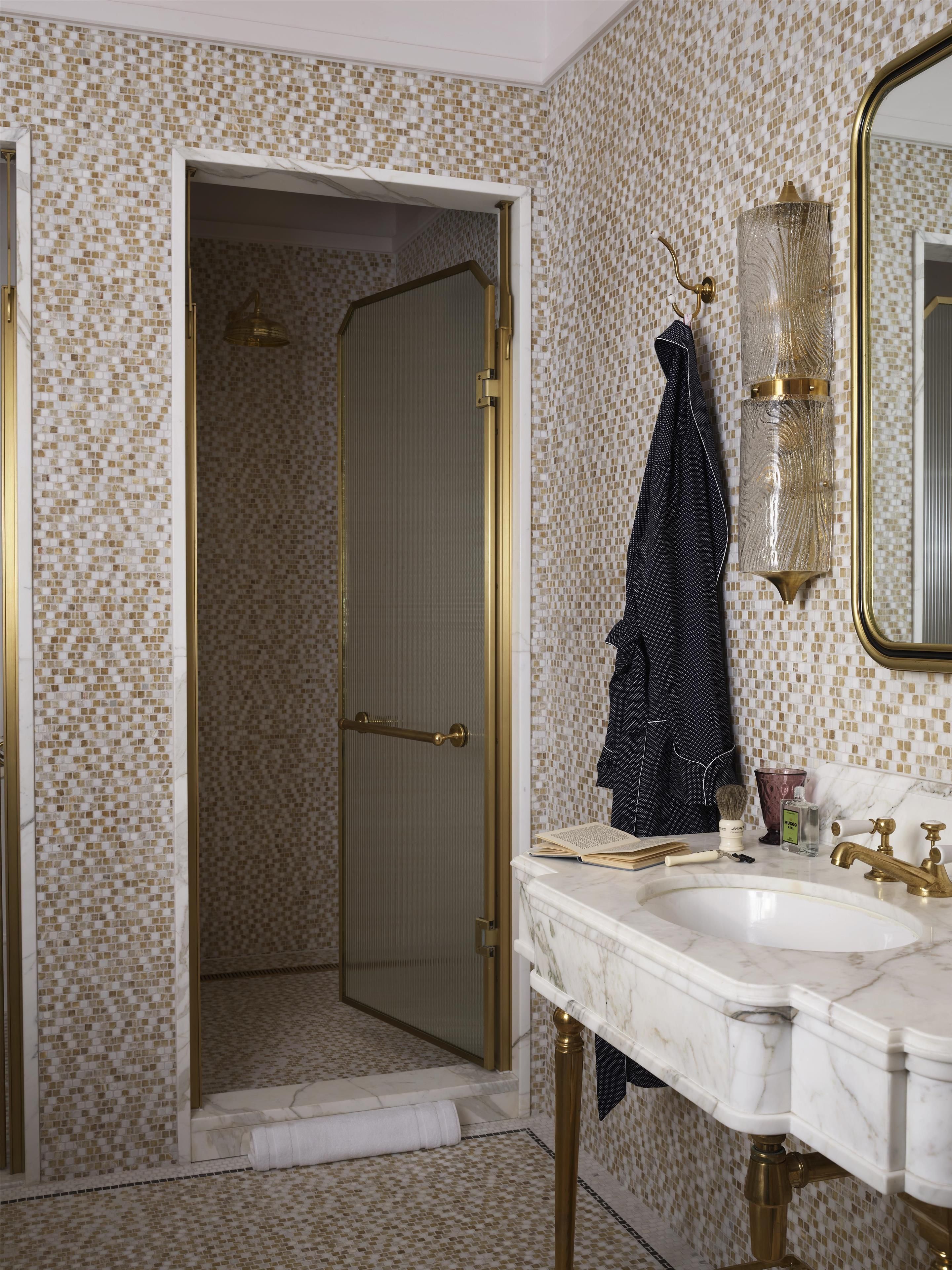 Un baño con lavamanos de mármol, grifería dorada, ducha con puerta de vidrio y pared con diseño.