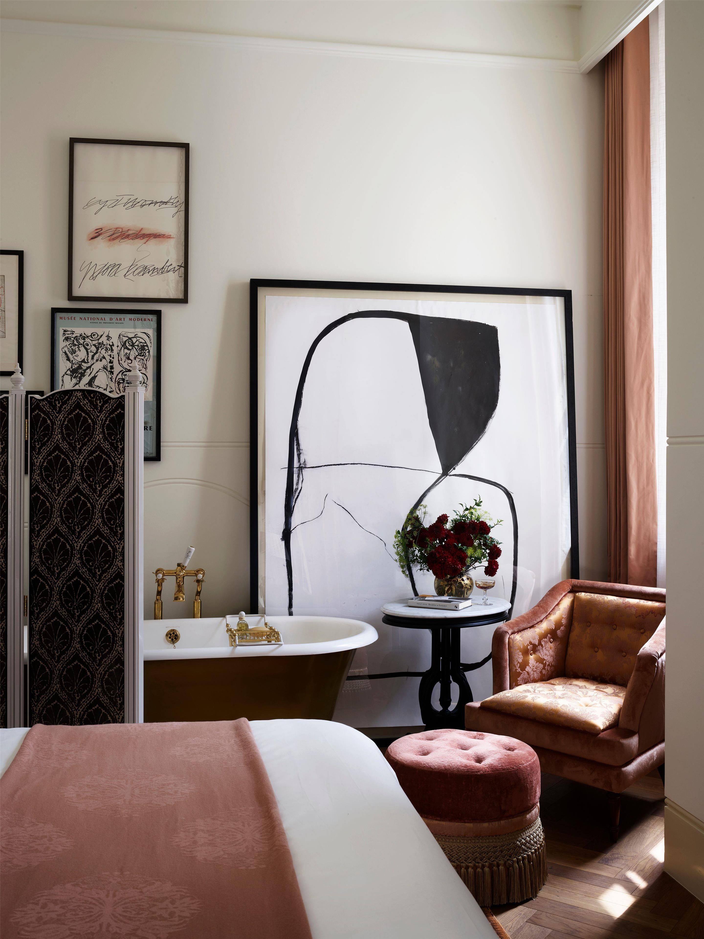 Et hotelværelse med en stor seng, et siddeområde med en lænestol og skammel, et badekar, et lille bord med en vase blomster, og et stort abstrakt maleri på væggen.