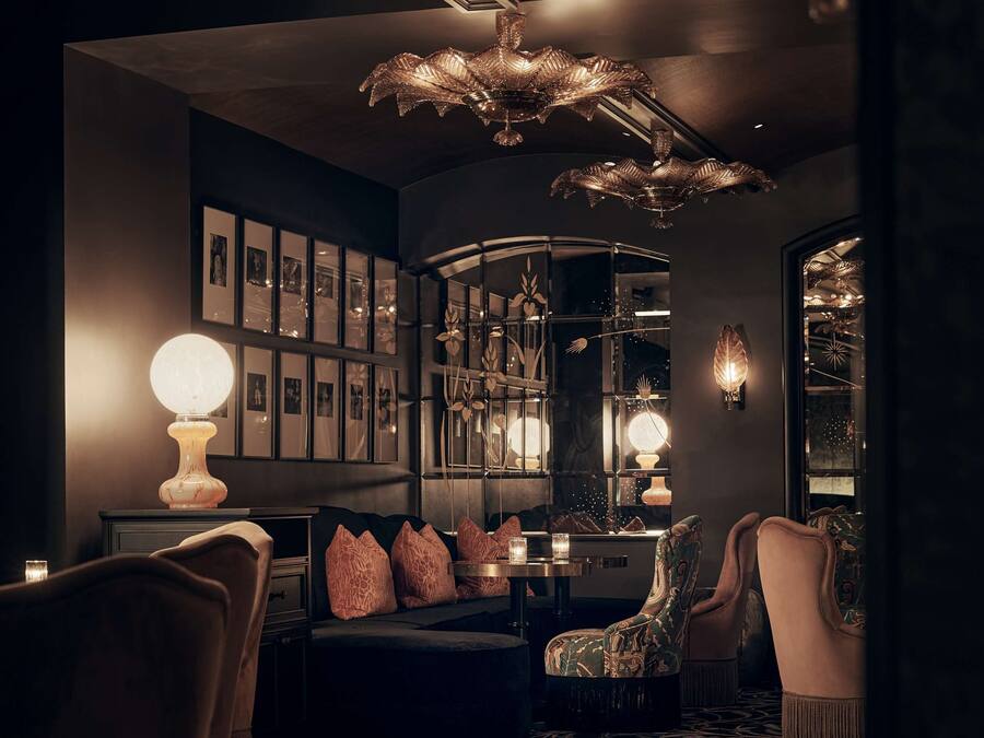 Uma área de estar com iluminação suave, tapete estampado, mesa redonda com abajur e disposição de assentos com cadeiras e sofá.