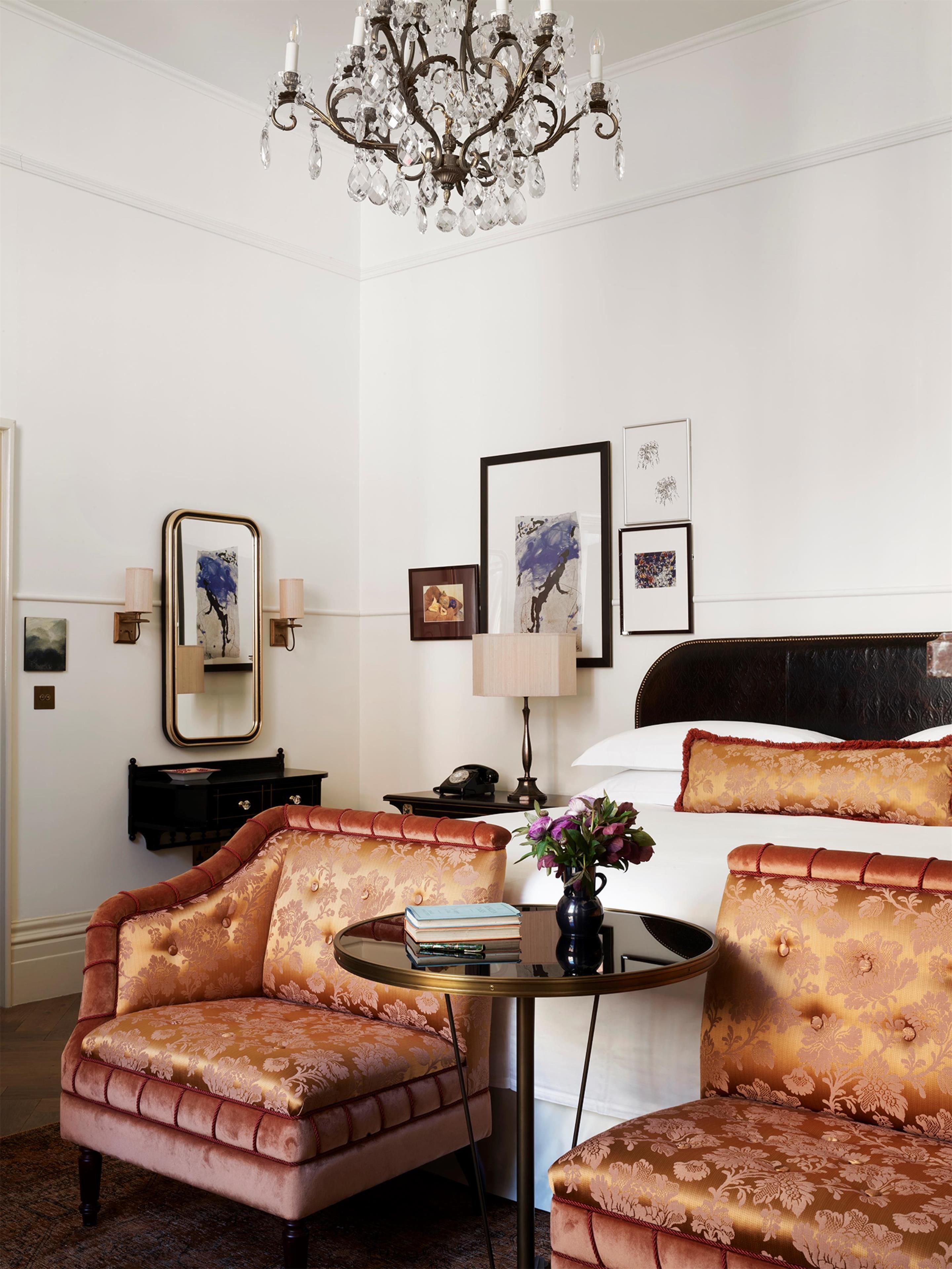 A hotel room with a bed, a sitting area with a chair and a table, a chandelier, and artwork on the wall.