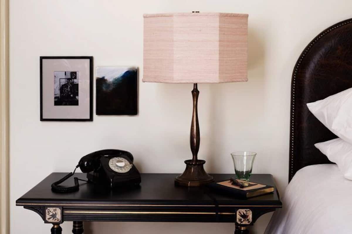 Uma cama bem arrumada com cabeceira escura, uma mesa de cabeceira com abajur, telefone e quadros na parede.