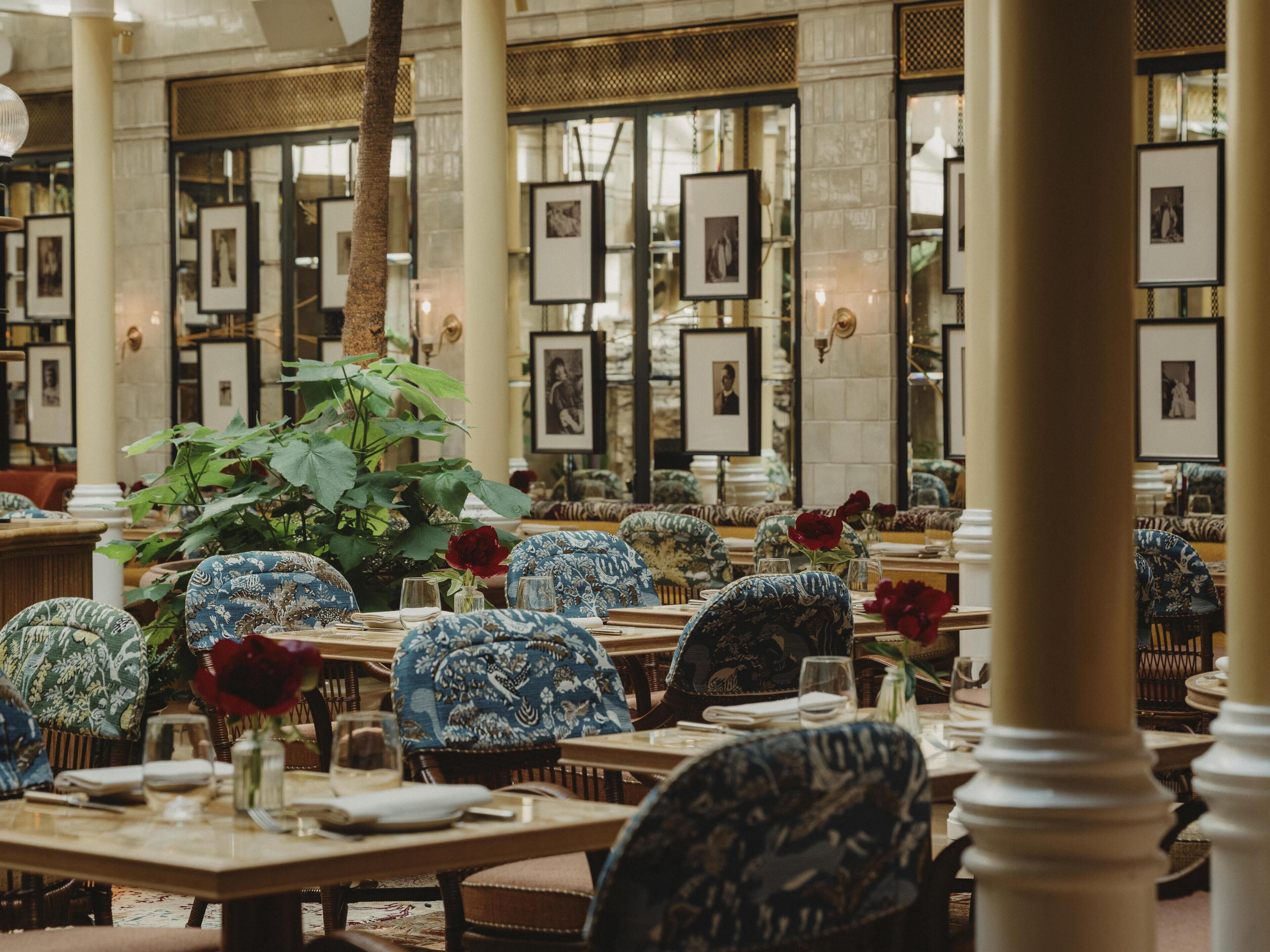 A dining area with tables set for a meal, framed pictures on the walls, and a large plant in the center.