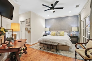 A bedroom with a bed, bedside tables, a desk, a chair, a ceiling fan, and a TV. - Inviting garden level apartment, The JAD Cox Suite (Savannah)