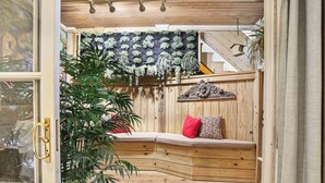 Un banc en bois avec des coussins, une plante suspendue et un tapis à motifs dans une pièce dotée d’un plafond en bois et d’un escalier.