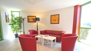 Una sala de estar con un sofá rojo, dos sillones rojos, una mesa de centro blanca y una lámpara de pie. Hay dos obras de arte enmarcadas en la pared y una planta en maceta.