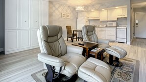 A modern living room with two leather armchairs, a wooden coffee table, and a dining area with a small table and chairs. A kitchen with white cabinets and appliances is visible in the background.