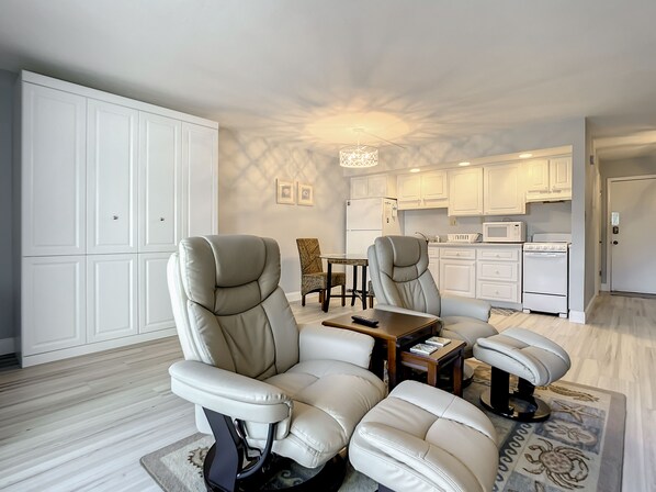 Una sala moderna con dos sillones de cuero, una mesa de centro de madera y un comedor con una pequeña mesa y sillas. Se ve una cocina con armarios y electrodomésticos blancos al fondo.