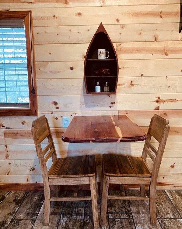 A wooden interior with a table and two chairs, a shelf with a mug and bottles, and a window with blinds.