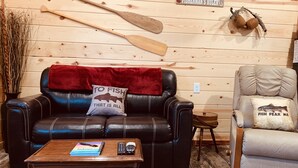 Une pièce chaleureuse avec un canapé en cuir, une table basse en bois et un fauteuil. On peut voir un mur en bois orné d’outils de pêche et d’une enseigne.