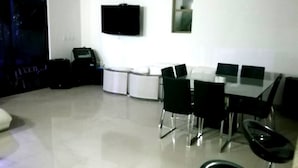 A modern dining area with a white table and black chairs, a flat-screen TV mounted on the wall, and a large window providing a view of the outside.