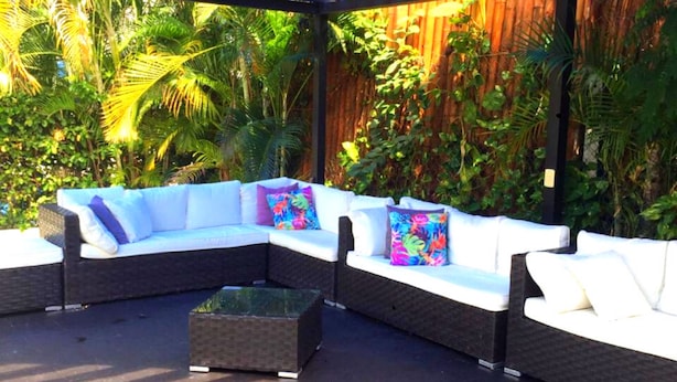 An outdoor seating area with a curved white sofa and cushions, a small square table, and a wooden fence in the background.