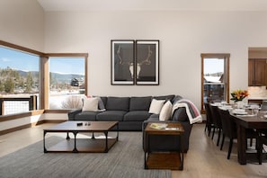 A modern living room with a large gray sofa, two framed antlers on the wall, a dining area with a wooden table and chairs, and a view of a snowy landscape through large windows. - 219 Elk Ranch Private Hot Tub (Fraser)