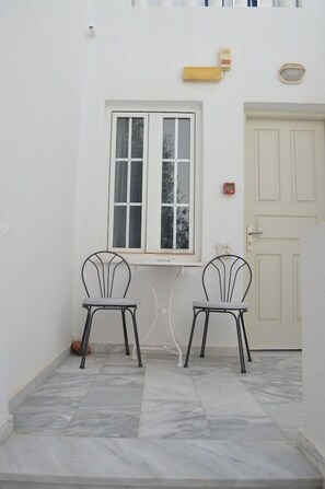 A white exterior wall with a window, two metal chairs, a door, and a small outdoor light fixture. - Kamari Beach (Santorini)