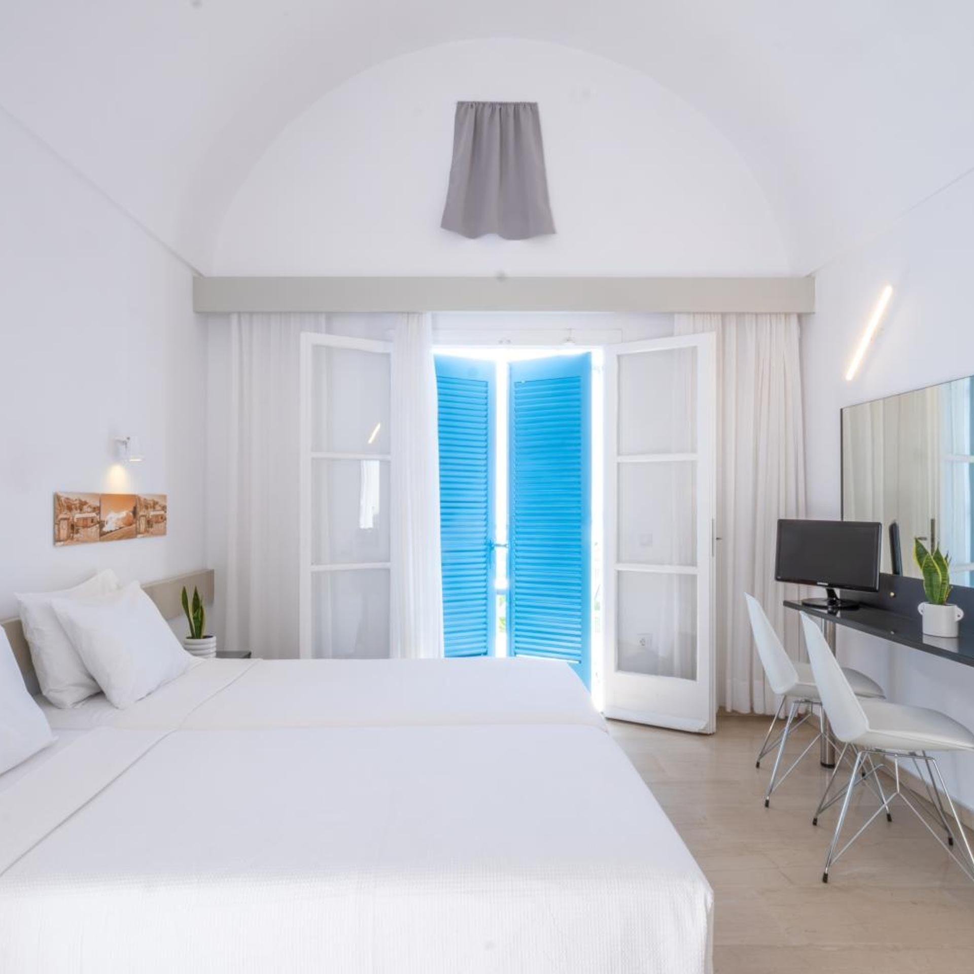 Habitación de hotel moderna con una cama grande, un escritorio con computadora y vista a través de una puerta con contraventanas azules.